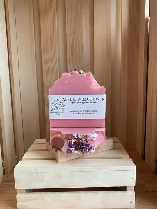 Alpine Wildflower handmade soap bars with pink color and dried flower pieces displayed on a wooden crate stand. 