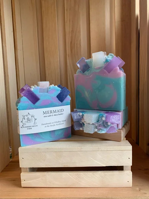 Mermaid handmade soap bars in pastel blue, purple, and teal colors displayed on a small wooden crate stand
