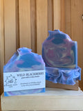 Two bars of Wild Blackberry goat milk and shea butter handmade soap bar with blue and purple swirl design on a wooden display. 