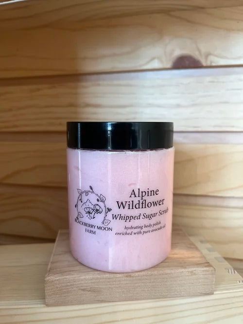 Alpine wildflower whipped sugar scrub body polish in a plastic jar with black lid displayed on a wooden stand. 