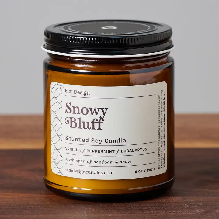 Snowy Bluff scented soy candle in amber glass jar with black lid on wood surface