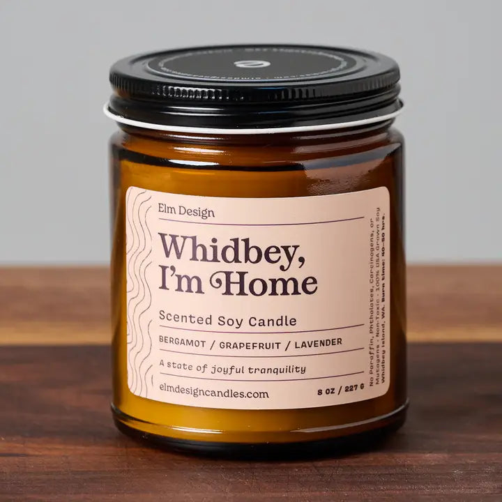 Whidbey, Im Home scented soy candle in amber glass jar with black lid on wood surface