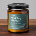 Whidbey Woods scented soy candle in amber glass jar with black lid on wood surface