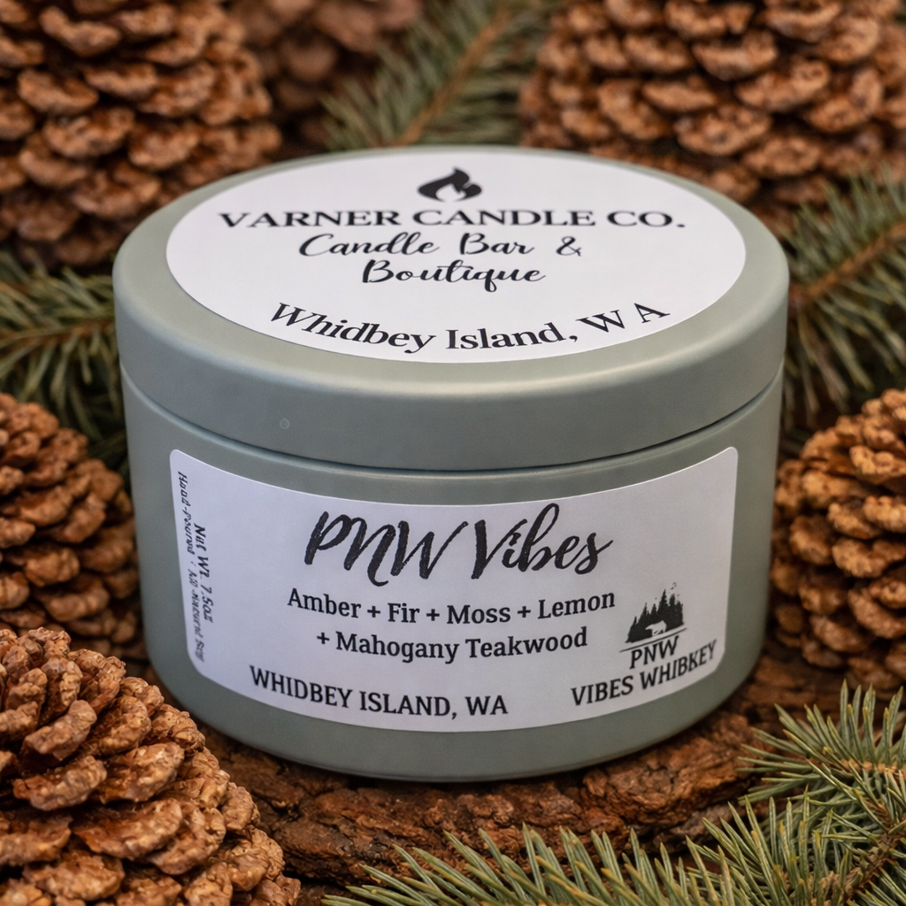 PNW Vibes scented candle in green tin labeled Amber, Fir, Moss, Lemon and Mahogany Teakwood on pinecone background
